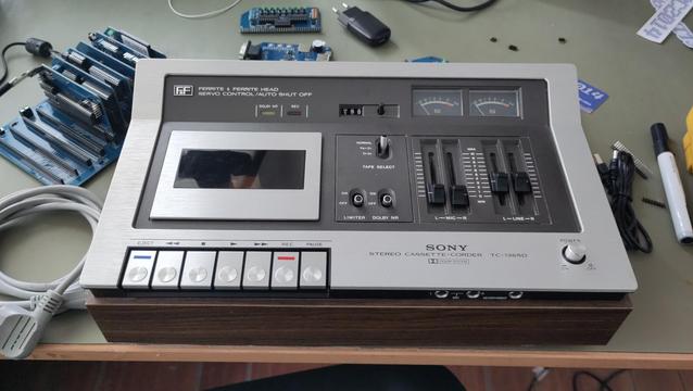 A silver and black top loading cassette tape deck with analog VU meters, knobs and sliders.