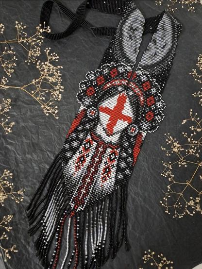 Traditional Ukrainian handmade beaded necklace. The decoration is made in black, red and white tones with a central symbolic image of a girl on her face made of a red cross. The lower part of the necklace is decorated with a long beaded fringe. The necklace is photographed on a dark background next to dry gypsophila twigs.