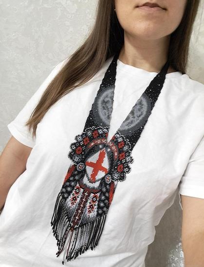 A photograph of a woman wearing a massive, long beaded necklace. The jewelry is handmade in traditional Ukrainian technique, with an ornament in black, white, red, and gray. The centerpiece of the necklace features a symbolic image of a red cross on a white background, surrounded by geometric patterns. The necklace has a long beaded fringe that falls onto a white T-shirt. The photo was taken from close range, from shoulders to chin, against a light wall.