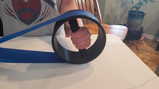 A hand gripping a black handle surrounded by a black wheel on a white countertop. There is a blue elastic band wrapped around the groove on the perimeter of the wheel to provide resistance when rotating.