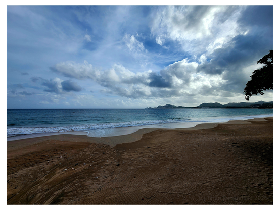 Wet sand etched with shallow channels leads into gentle surf beneath a vast sky crowded with sculpted clouds. A chain of green hills traces the distant coastline as the sea shifts from turquoise to deep blue. The beach is empty, and the changing light gives the scene a sense of calm just before or after passing weather.