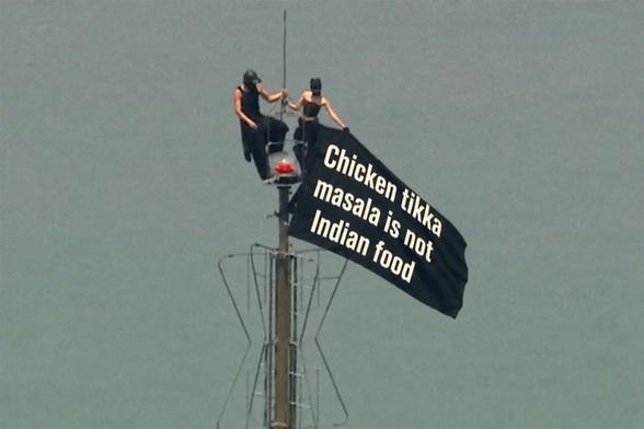 The Empire State climbing couple unfurl a black flag but it reads “Chicken tikka masala is not Indian food”