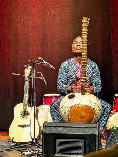 Ein Mann in traditioneller Kleidung spielt auf einer Kora, einem westafrikanischen Instrument, auf einer Bühne. Daneben stehen eine Akustikgitarre und mehrere Trommeln.