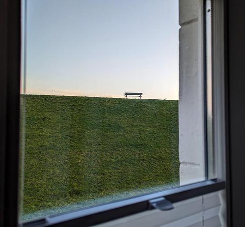 Blick durch ein Fenster auf eine einsame Parkbank auf einem grasbewachsenen Deich bei klarem Himmel.