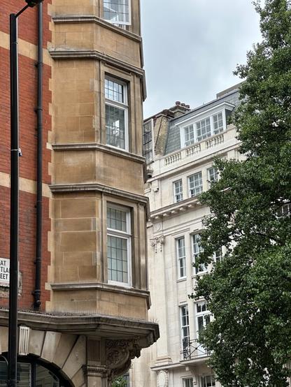a multi-storey octagonal stone bay emerging at the corner of a brick building, above the ground floor. Along the side facing the street are windows, each divided into multiple panes. In the background, a white stone building can be seen.