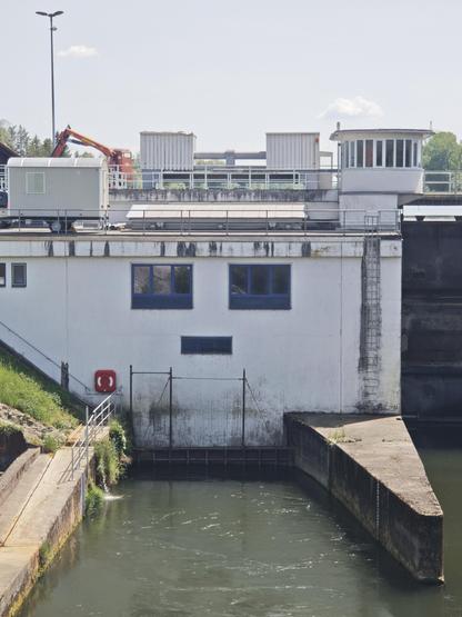Das Foto zeigt ein weißes Industriegebäude an einer Wasserstraße, das optisch an ein Gesicht erinnert. Eine weiße Betonwand mit zwei blauen Fenstern (Augen) und einem Lüftungsschlitz (Mund). Im Vordergrund trübes, fließendes Wasser mit einer Betonmole auf der rechten Seite, die mit dem Betonteil links ein wenig wie geöffnete Arme aussehen. Im Hintergrund auf dem Dach befinden sich ein Laternenmast, Container, ein Anhänger, ein kleiner Kontrollturm und ein orangefarbener Kranarm. Auf der linken Seite unten ein grüner Hang mit einer Treppe und einem roten Rettungsring an der Betonwand.