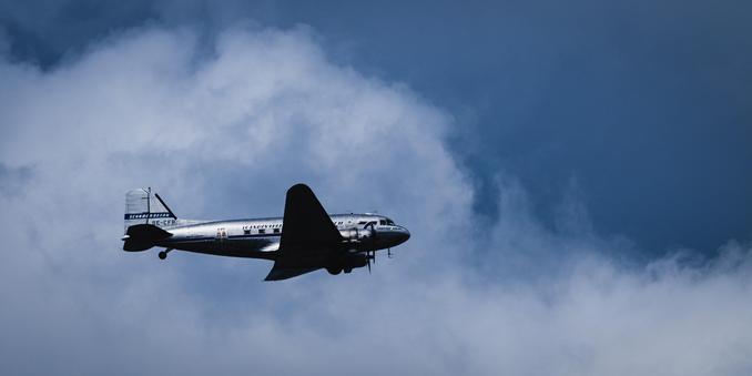 A side profile panoramic format contrasty photograph of a Douglas DC-3 propeller passenger airplane, made in 1943.

The metallic airplane, underside in almost black shadow is flying to the left and with blue stripes painted along the side of the body and tail, is flying in front of a fluffy white cloud formation that occupies the lower half of the photo. The upper half is mostly blue sky.
