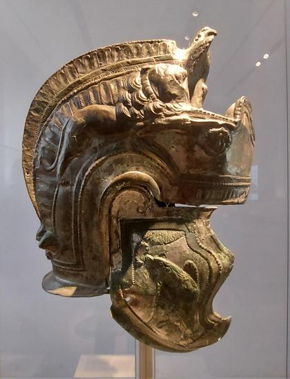 A highly decorated helmet made, featuring intricate reliefs of animals such as a lion and an eagle, ornate patterns along the crest, and detailed engravings on the cheek guard, displayed against a neutral background