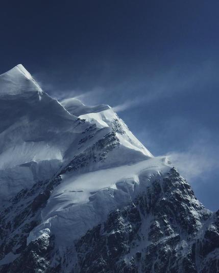 Close-up photograph of Aoraki / Mount Cook, New Zealand, showing snow-covered peaks and dark rocky ridges. Strong winds blow snow from the summit beneath a deep blue sky, highlighting the mountain's dramatic alpine conditions.