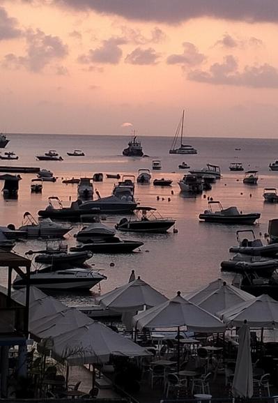 the suns first breach, a small slither of sunlight appears just at the horizon, the sea and small boats are bathed in the orange light as the sun places its first kiss onto the sky and the sea. everything is thrown into silhouette, except white umbrella canopy's and the white of the hulls of the numerous small boats in Xemxija Bay