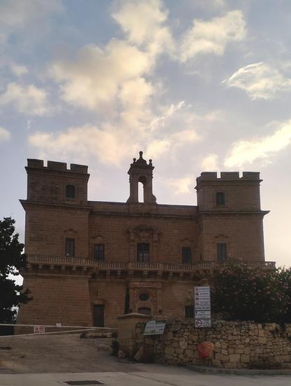 the image is of a castle like structure, known as Selmun Tower (also known as Selmun Palace) it is a limestone block structure set in a small fenced off court. wispy white clouds pass overhead in a bright bluw sky