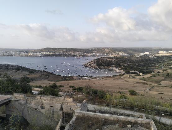 View from the site of the former Selmun Fort, The buildings have since been fenced off as they are dangerous and falling to pieces, however the view from the site of the fort looks back over Mistra Bay and then around the headland on the right Xemxija Bay. Over the water, apartment blocks on the sea front at St Pauls Bay line the coast of the Mediterranean sea. Hills and countryside form an in land horizon, where land and sky meet it is a hazy blue, the sky filled with wispy clouds