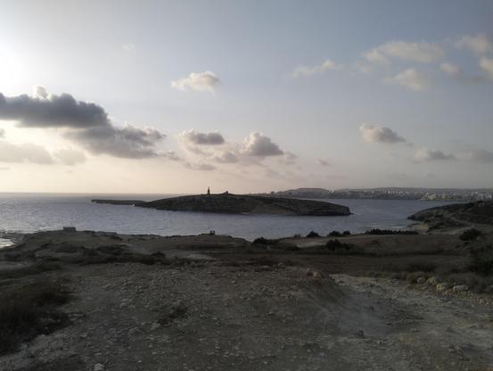 A wide angle view of St Pauls Island from blata il-bajda, the stone cross visible on the island, beyond tha forming the horizon is St Pauls Bay. The coast line snakes around from blata il-bayda to Mistra bay and then to Xemxija Bay and to St Pauls Bay. In the immediate foreground the trail goes off in multiple directions all along a stony dusty ground