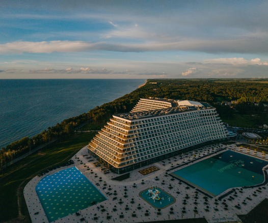 Hotel Gołębiewski
A concrete structure, besides a shoreline, in the shape of a pyramid with the top cut off stretched along a symmetrical “L” shape. Behind it there is some greens with forests or parks, inf front a concrete-laid area with single small trees or bushes, something that looks like pools or artificial greenery, no shade in sight.