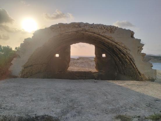 An artillery post -  made of an iron lattice that is slowly rusting away and covered with a natural limestone top to blend it in with the rest of the gnarly rocks in this area, most likely used in World war two . The sun is just above and to the right as a bright white yellow orb in a blue cloudy sky.