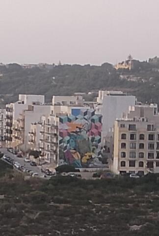 View looking down from the road leading upto the selmun tower, a white apartment block in Xemxija has been brightly painted on one side with green, grey, red and yellow cubes in 3D