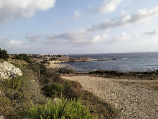 Looking back along the coastal trail towards Blata il-Bajda from Mistra bay. A stony trail is lined with heather on the left side and a rocky coast line with salt pans just visible on the right. The sky is blue and filled with fluffy white clouds