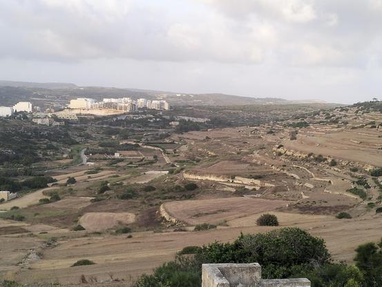 View of the Xemxija / Mistra / Selmun countryside. White buildings of Xemxjia are visible in the distance, but the foreground is made of farmers fields arranged in steps to maximise the amount of growing area available on a slope. At the top of the hill on the right Selmun tower sits (though not pictured. The hill runs all the way down to Mistra bay and then out and up to the outskirts of Xemxija