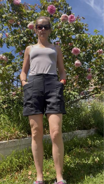 A woman in a grey tank top and black shorts stands in front of a flowering rose bush. The shorts are mid-thigh length with a fly zip, large angled pockets, and a turned up cuff - front view.