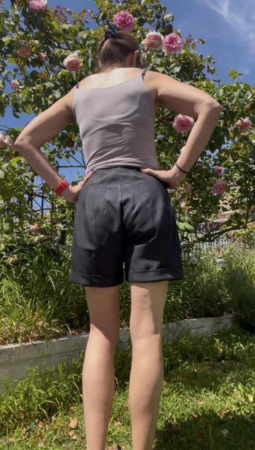 A woman in a grey tank top and black shorts stands in front of a flowering rose bush. The shorts are mid-thigh length with rear darts, large angled pockets, and a turned up cuff - back view.