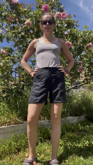 A woman in a grey tank top and black shorts stands in front of a flowering rose bush. The shorts are mid-thigh length with a fly zip, large angled pockets, and a turned up cuff - front view.