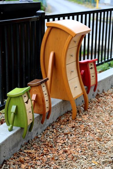 Four curved wooden cabinets in bright colors outside sitting on a kerb, three seemingly chipper and smiling. A larger one looking tired, slightly hunched forward.