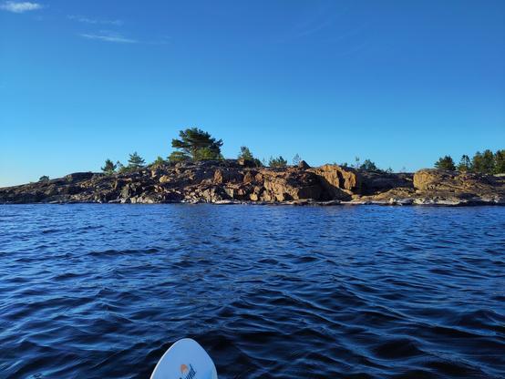 A rocky shore between blue water and blue skies, the shapes square and jagged. Behind the shore there are a couple of trees. Half a paddle is in the lower frame.