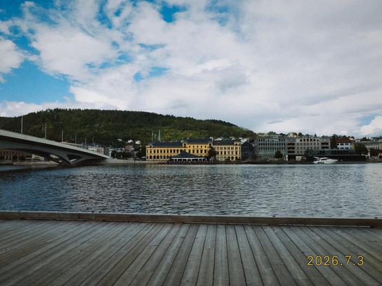 Matt is sitting with his back to a train station and his front to a body of water. To the left the bridge crosses from one side of the water to the other  and review on the other side is lined with short buildings and hillside.