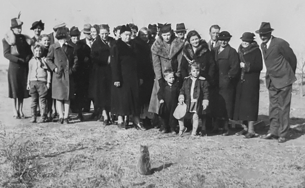 Black-and-white photo of a couple dozen light skinned people all wearing dressed up outdoor clothing. They are all standing outside in a scrubby field as of about to pose for a group portrait, but they’re all looking down at a small tabby cat that sits right in front of the group and looks calmly back at them.