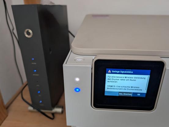 Ein weißer Multifunktionsdrucker steht neben einem schwarzen WLAN-Router auf einer Holzoberfläche. Das Display des Druckers zeigt eine deutsche Warnmeldung "Geringe Signalstärke" mit der Empfehlung, den Drucker näher am Router aufzustellen für eine bessere Wireless-Verbindung. Blaue LED-Lichter blinken am Router und Drucker.