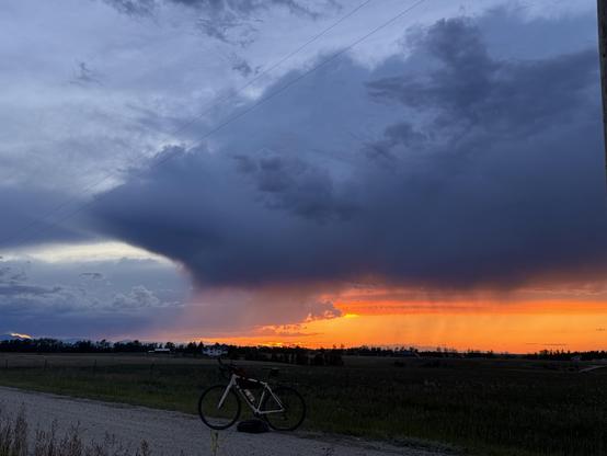 A thunder cloud at sunset seen across the East Bench north of Red Lodge Montana. A white gravel bike is parked on a gravel road in the foreground. The cloud is trailing rain lit red and orange by the sunset bringing much needed moisture after a warm dry winter.