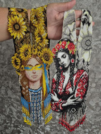 The hand holds two long. intricate Ukrainian gerdans (beaded jewelry), each decorated with a beaded portrait of a Ukrainian woman. The left one, with vellow and blue hues. features sunflowers and a woman wearing patriotic glasses. The right one, in white, black, and red, shows a woman in traditional dress. Both have beaded fringe.