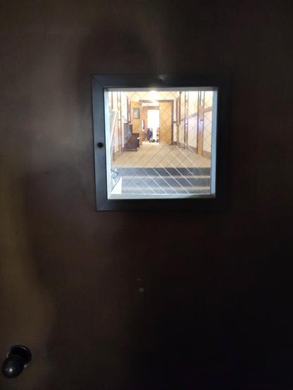 Photo taken inside a church. A small square window in an interiorcdoor shows a glimpse down a hallway. My shadow is visible on the door.