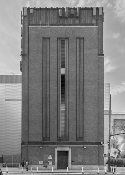 A roughly six story simple industrial art deco-style building, lacking windows, but with a narrow louvered vent at center running vertically along its length, across a city street.