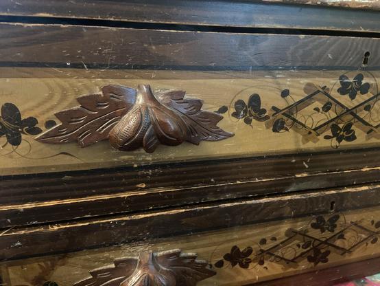A picture of the drawers of an old dresser. The handles are made to look like figs(?) or walnuts I have no idea as the leaves are wrong and in fact as a kid they looked like two funny textured breasts 😹
The whole surface is hand painted with brown stripes to make it look like wood grain (in two tones one set is dark chocolate brown (two shades of that for the grain) and the other a caramel brown in the “center” (also two shades for the grain). There is a painted “framing” around the caramel brown and splotchy painted leaves with tendrils on the sides and around a sort of painted “lattice”