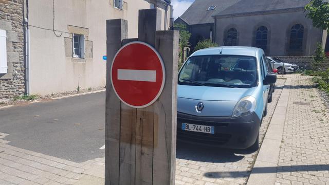 Une rue avec un trottoir pavé.
Au premier plan, un panneau sens interdit sur un support en bastaings.
Derrière ce anneau, une voiture bleue ciel garée.
Sous le panneau, on distingue une forme plus claire rectangulaire, marquant un panneau qui vient d'être retiré.