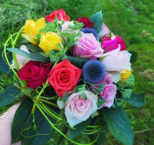 Ein farbenfroher Blumenstrauß mit verschiedenen Rosen und blauen  Distelblüten wird von einer Hand gehalten. Der Strauß enthält rote, gelbe, rosa sowie hellrosa Rosen und blaue, kugelförmige Blüten. Grüne Blätter und dünne, gewundene Ranken sind zwischen den Blüten angeordnet. Im Hintergrund ist unscharfes grünes Gras zu erkennen.