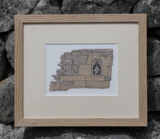 Framed stitched artwork depicting a small multipaned arched window set in a sandstone wall