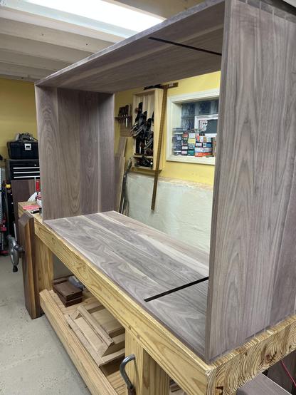 A large cabinet case made of walnut wood. It is on its side on top of a woodworking bench.