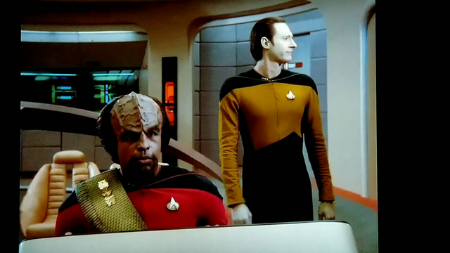 Star Trek The Next Generation scene, and it's a VIDEO! OK, we're on the bridge and looking at Mr. Worf in red uniform sitting at a console in those cool lean-back pleather chairs. Standing behind him alone is Data, in gold uniform. Data's standing casually and looking to his left and slightly nodding like he's in convo with someone. Then, he rests his elbow on a nonexistent something and leans on it and falls immediately over to the floor because the thing he leaned on is not really there. Heh. Data's doing bits.