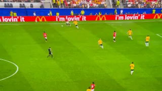A World Cup soccer game with the animated ad screen behind the game being played. A giant 50-foot purple Grimace pops up in the thin strip of the video screen. Only his eyes and parts of his waving hand are visable.