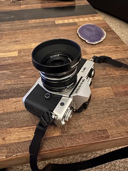 An OM System OM-3 camera is seen on a coffee table made of varying colored wood segments. It’s a mix of black textured plastic and silvery metal with lots of buttons and dials.  The lens has a round metal lens hood attached. The lens is pointing up with the body flat on its back. The lens and hood markings say “Olympus” because the lens is 10 years old.