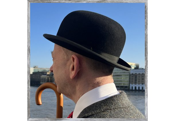 head shot - Scott Rochester is standing on London Bridge looking away from the photographer towards Tower Bridge - he is wearing a jacket and tie, and a bowler hat - he is carrying an umbrella on a bright sunny day without a cloud in the sky (this is his Patrick Macnee impression) - in the middle distance is the River Thames - beyond that the buildings of the South Bank including the London Bridge Hospital and the Hays Galleria