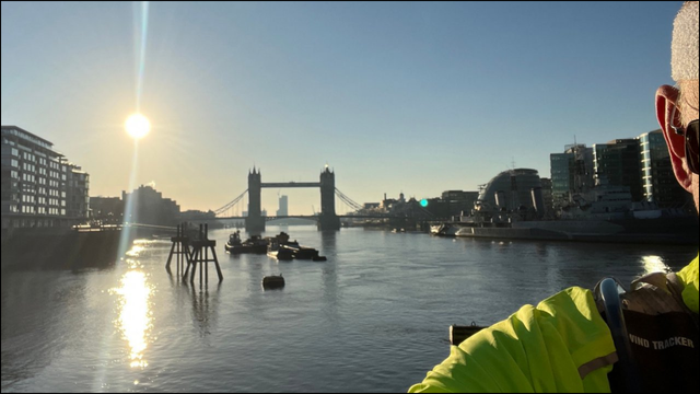 A photograph of the River Thames and Tower Bridge at sunrise with a perfect blue sky. Taken at 7.21am on Sunday 16 March 2025. The Sun is up and dominates the photo just above the buildings to the left of the bridge. Scott Rochester is standing to the right hand side of the photo, deliberately hiding his face. He has a dayglo yellow running top, a runners vest with a water supply, tinted reactolite glasses, and white terylene sweatband on his head.