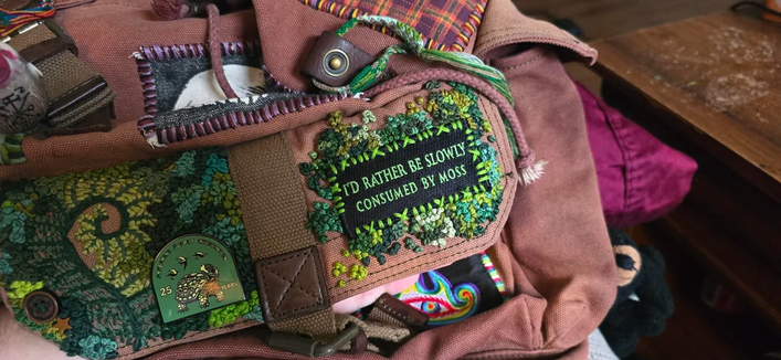 A heavily embroidered bag with moss and fern motif. Also includes a patch that says "I'd rather be slowly consumed by moss" and a pin with a turtle on it.