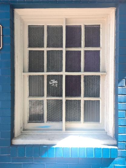 White muntined window in a blue brick wall. There are sixteen panes of privacy glass, in slightly varying colors. Heavy muntins divide the window in quarters and thinner ones divide each quarter into quarters again. One pane has script graffiti in white paint "Tech Destroys"