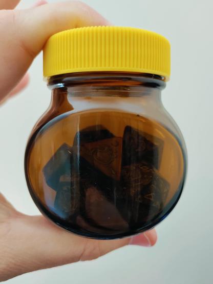 A hand holds a small brown glass bottle with a yellow lid. The silhouette of a set of polyhedral dice can be seen through the glass.
