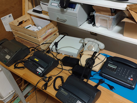 Foto einer Holz-Werkbank auf der mehrere Rollenfaxgeräte stehen, Wählscheiben-Telefone und eine alte ISDN-Telefonanlage mit analog-Anschlüssen.