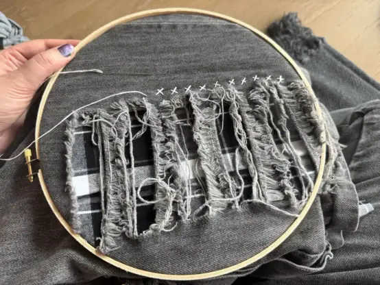 An embroidery hoop containing a section of a pair of grey wash denim Tripps. The section has a fashionable ladder rip in it; a piece of black and white plaid flannel has been placed behind the rips and a hand is in the process of sewing the two pieces of fabric together with white dental floss in a pattern of small Xes.