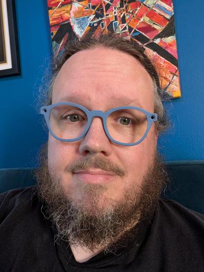 Fish sitting in front of a deep blue wall with some brightly colored artwork hanging from it. He is wearing a fairly normal set of eyeglasses, though you might notice that the front texture is that of a build plate from an FDM printer.